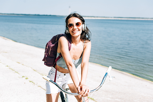 smiling girl with backpack biking and listening music in headphones near river in summer