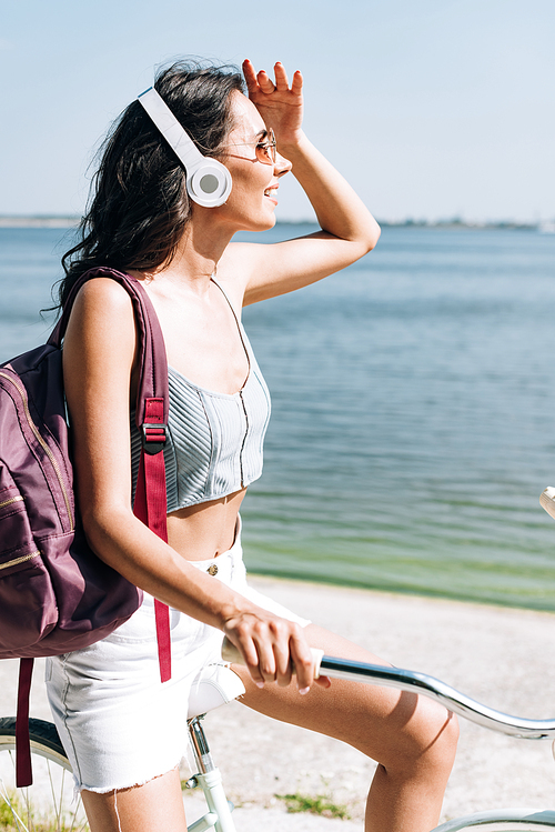 happy brunette girl with backpack biking and listening music in headphones near river