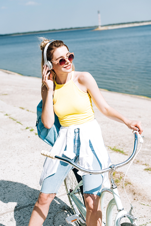 blonde girl with backpack biking and listening music in headphones near river in summer