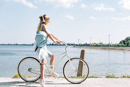 side view of girl with backpack biking and listening music in headphones near river in summer