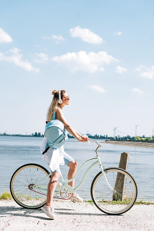 side view of girl with backpack biking in headphones near river in summer