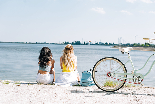 back view of blonde and brunette friends sitting near bike and river in summer
