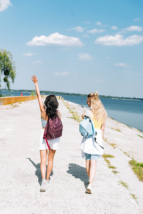 back view of blonde and brunette girls walking with backpacks near river in summer