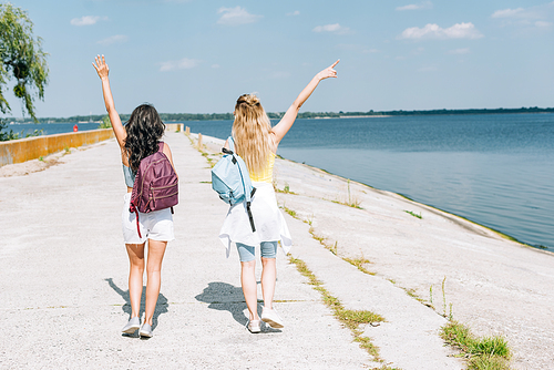 back view of blonde and brunette girls walking with backpacks and hands in air near river in summer