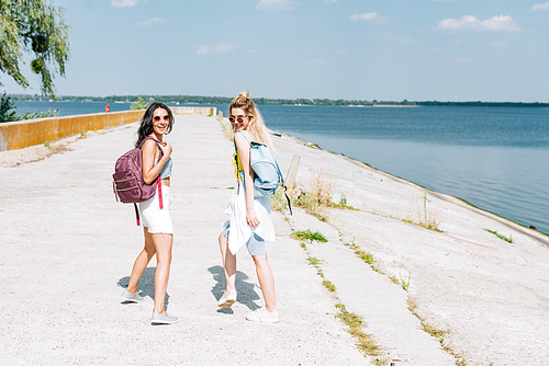 happy blonde and brunette girls walking with backpacks near river in summer