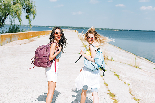 cheerful blonde and brunette girls walking with backpacks near river in summer