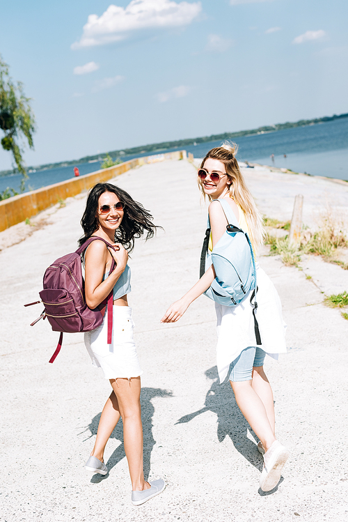 cheerful girls walking with backpacks near river in summer