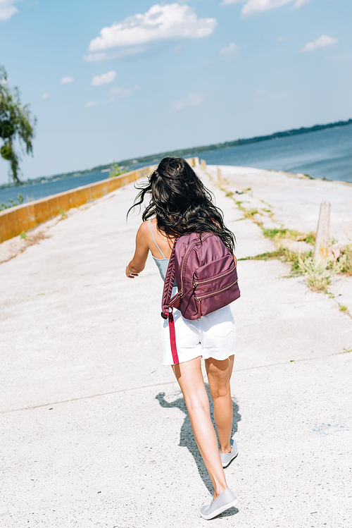 back view of brunette girl walking with backpack near river in summer