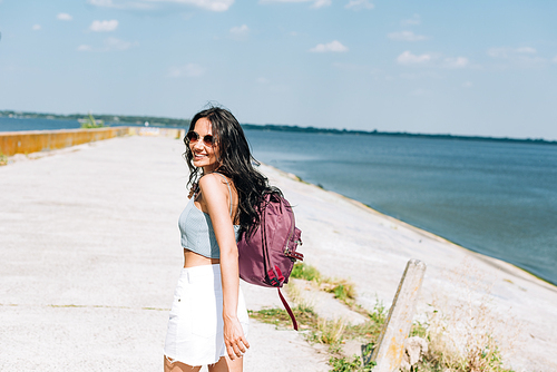happy brunette girl walking with backpack near river in summer