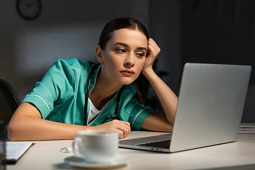 attractive nurse in uniform sitting at table and looking at laptop during night shift