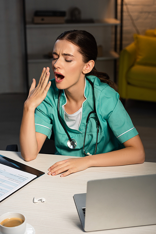 attractive nurse in uniform sitting at table and yawning during night shift