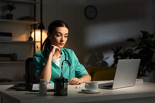 attractive nurse in uniform sitting at table and looking at laptop during night shift