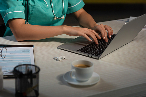 cropped view of nurse in uniform sitting at table and using laptop during night shift
