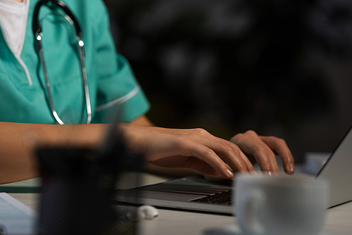 cropped view of nurse in uniform sitting at table and using laptop during night shift
