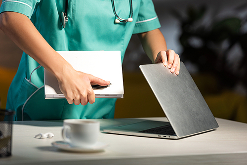 cropped view of nurse in uniform holding notebook and closing laptop during night shift