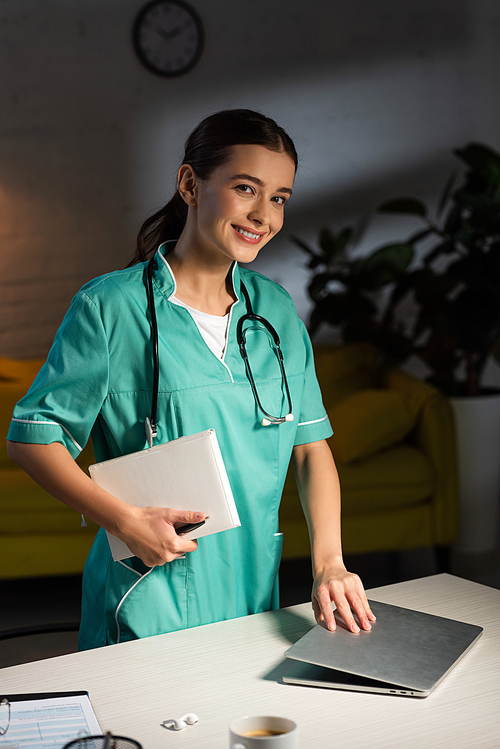 smiling nurse in uniform holding notebook and closing laptop during night shift