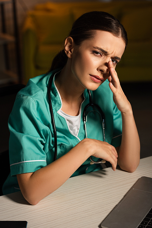attractive and tired nurse in uniform sitting at table and crying during night shift