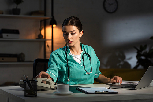 attractive nurse in uniform sitting at table and taking handset during night shift