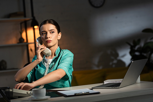 attractive nurse in uniform sitting at table and talking on telephone during night shift