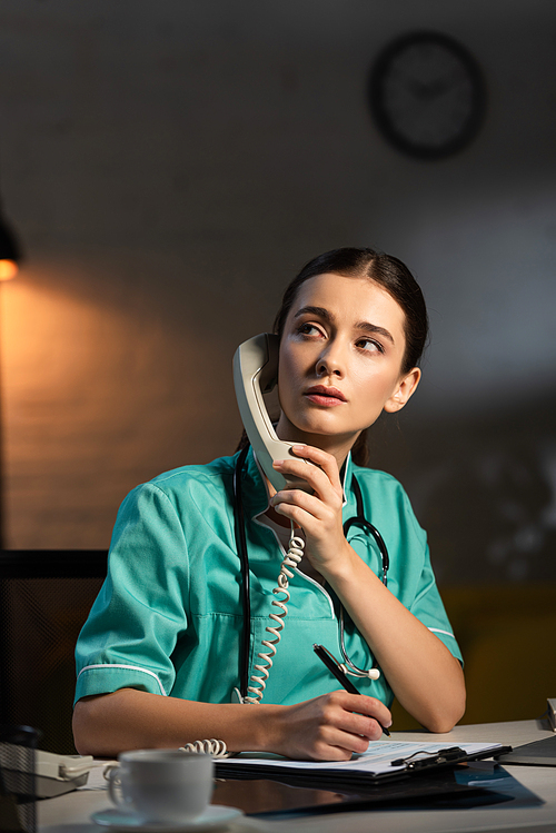 attractive nurse in uniform sitting at table and talking on telephone during night shift
