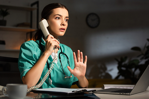 shocked nurse in uniform sitting at table and talking on telephone during night shift