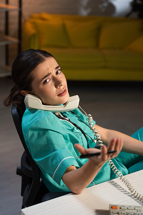 attractive nurse in uniform sitting at table and talking on telephone during night shift