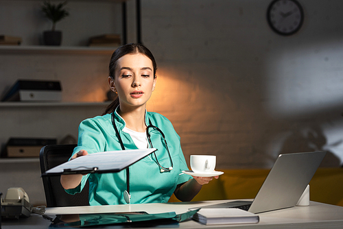 selective focus of attractive nurse in uniform holding clipboard and cup during night shift
