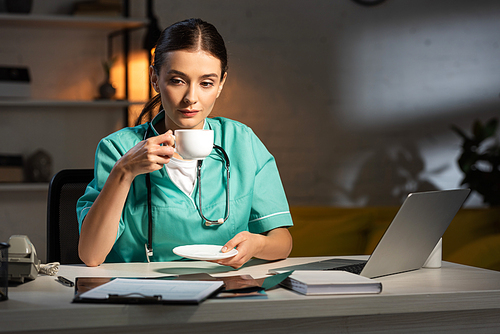 attractive nurse in uniform sitting at table and drinking coffee during night shift
