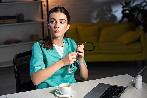 attractive nurse in uniform sitting at table and holding stethoscope during night shift