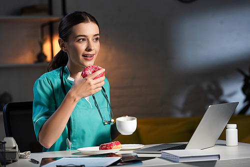 attractive nurse in uniform sitting at table and eating donut during night shift