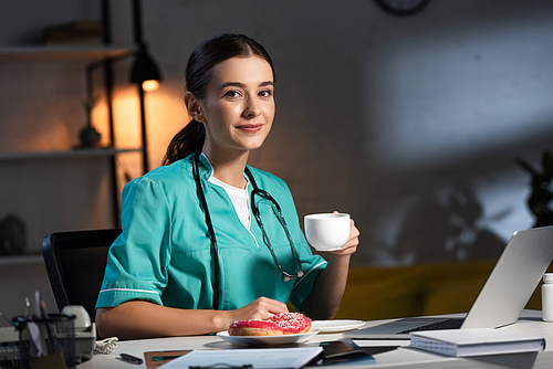smiling nurse in uniform sitting at table and holding cup during night shift