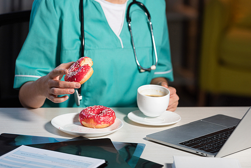 cropped view of of nurse in uniform holding donut and cup during night shift