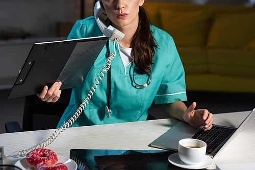cropped view of nurse in uniform talking on telephone and holding clipboard during night shift