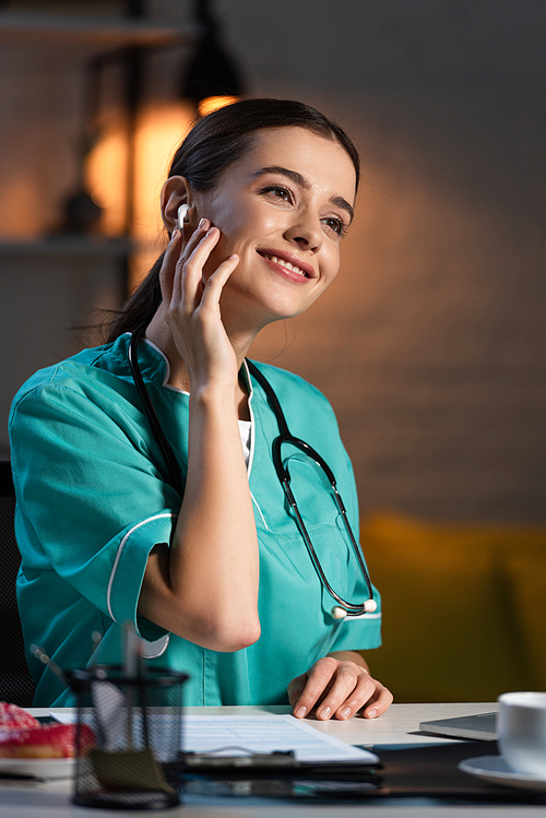 attractive and smiling nurse in uniform sitting at table and listening music during night shift
