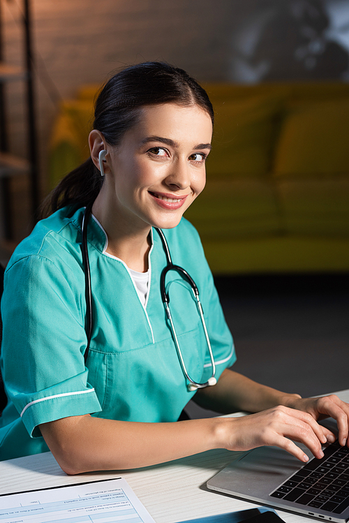 attractive and smiling nurse in uniform using laptop and listening music during night shift