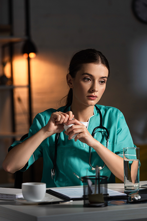 attractive nurse in uniform sitting at table and opening bottle with pills during night shift