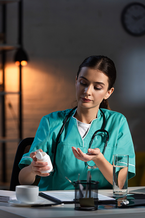 attractive nurse in uniform sitting at table and taking pill during night shift
