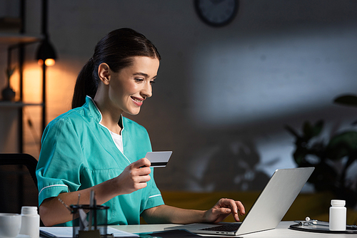 smiling nurse in uniform nurse in uniform holding credit card and using laptop during night shift