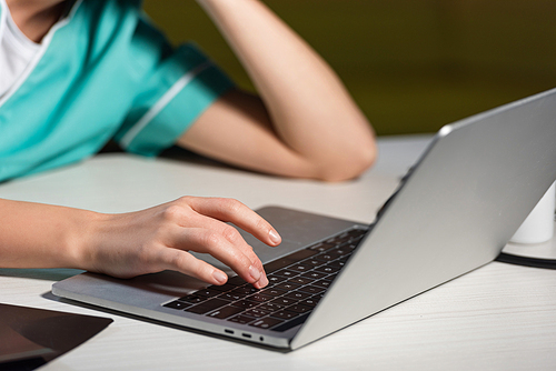 cropped view of nurse in uniform sitting at table and using laptop during night shift