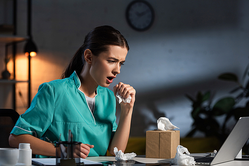 attractive nurse in uniform sneezing and holding napkin during night shift