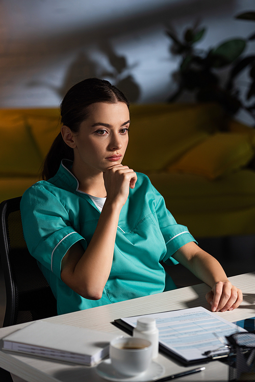 attractive and pensive nurse in uniform sitting at table during night shift