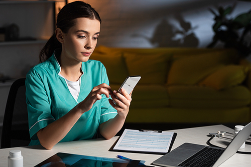 attractive nurse in uniform sitting at table and using smartphone during night shift