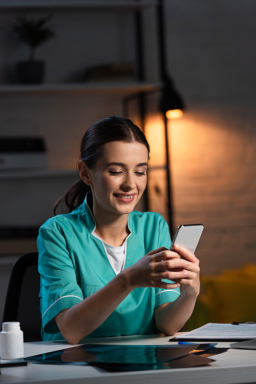 smiling nurse in uniform sitting at table and using smartphone during night shift