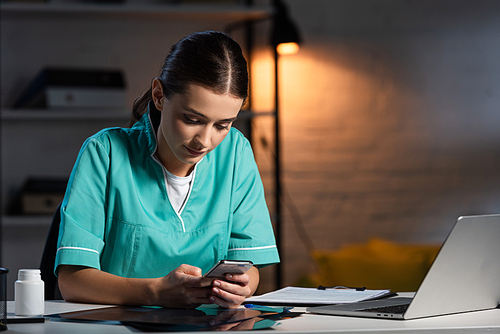 attractive nurse in uniform sitting at table and using smartphone during night shift