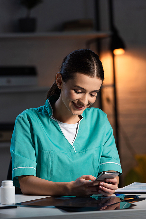 attractive nurse in uniform sitting at table and using smartphone during night shift