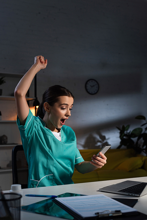 shocked nurse in uniform showing yes gesture and using smartphone during night shift