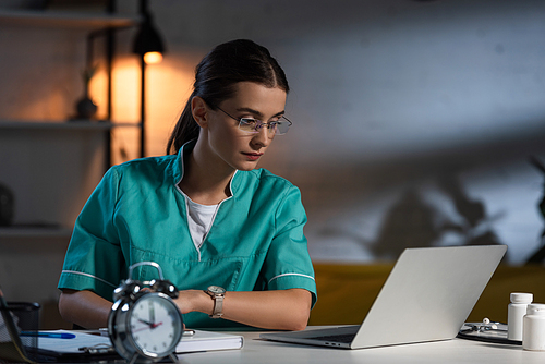attractive nurse in uniform and glasses sitting at table and looking at laptop during night shift