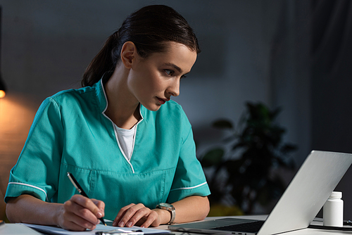 attractive nurse in uniform sitting at table and writing during night shift