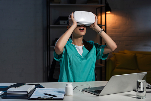 shocked nurse in uniform with virtual reality headset sitting at table during night shift