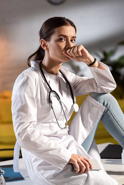 attractive doctor in white coat with stethoscope holding smartphone during night shift